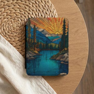 May include: A rectangular case featuring a vibrant landscape design. The artwork depicts a lake surrounded by mountains and trees, with a setting sun radiating golden light. The color palette includes blues, greens, and oranges.