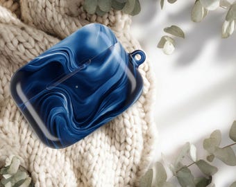 May include: A blue and white marbled AirPod case rests on a cream-colored knitted blanket. The case has a glossy finish and a small loop for attaching a lanyard. Eucalyptus leaves are in the background.
