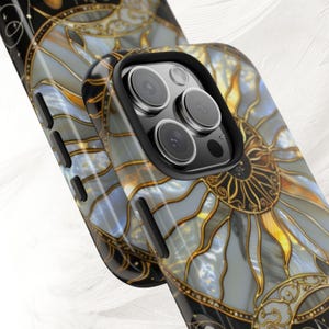 May include: Close-up of two phone cases with a stained-glass sun design. The cases feature a black border and a detailed pattern in shades of gray, gold, and blue. The design resembles a sunburst with intricate lines and shapes.