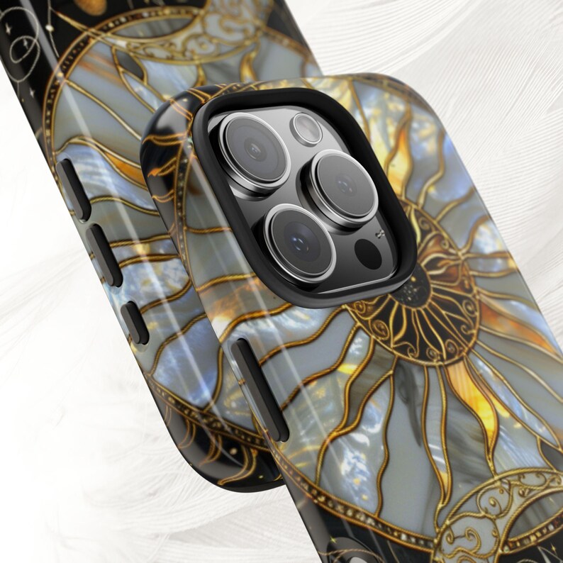 May include: Close-up of two phone cases with a stained-glass sun design. The cases feature a black border and a detailed pattern in shades of gray, gold, and blue. The design resembles a sunburst with intricate lines and shapes.