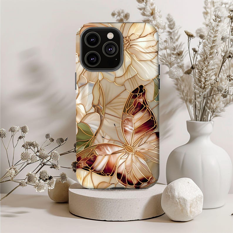 May include: A phone case with a stained glass design featuring a butterfly and flowers in shades of brown, cream, and gold. The case has a glossy finish and a raised camera bump.