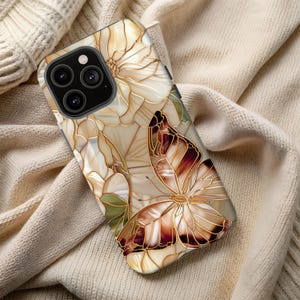 May include: A phone case featuring a butterfly and floral design. The case has a beige background with a stained glass effect. The butterfly has brown and white wings with gold outlines. The flowers are white with gold outlines.