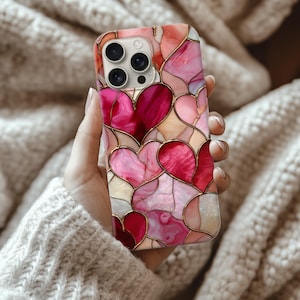 Puede incluir: Funda de teléfono con diseño de vidriera con formas de corazón en tonos rosa y rojo, contorneadas en dorado. La funda tiene un acabado brillante y un diseño protector.