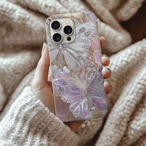 May include: A decorative phone case featuring a butterfly design with white and lavender wings, embellished with sparkling rhinestones. The case has a pearlescent background with pink and white accents, creating a glamorous look.