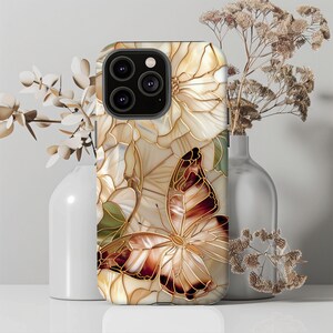 May include: A phone case featuring a butterfly and floral design. The case has a cream-colored background with a gold outline. The butterfly has brown and gold wings. The case is decorated with white flowers and green leaves.