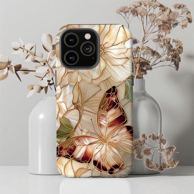 May include: A phone case featuring a butterfly and floral design. The case has a cream-colored background with a gold outline. The butterfly has brown and gold wings. The case is decorated with white flowers and green leaves.