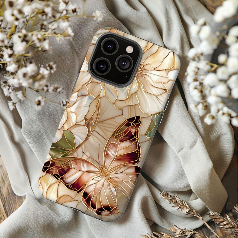 May include: A smartphone case featuring a butterfly and floral design. The case has a cream-colored background with a stained-glass effect. The butterfly has brown and red wings, and the flowers are outlined in gold. The case is designed for a smartphone.