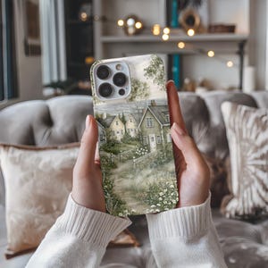 May include: A smartphone case featuring a detailed illustration of a coastal village with yellow and green houses, a white picket fence, and a garden path surrounded by green foliage and small white flowers under a soft sky.