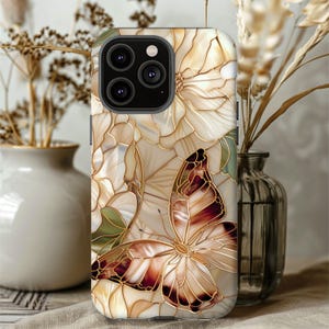 May include: A phone case featuring a butterfly and floral design. The case has a cream-colored background with gold-colored outlines. The butterfly has brown and cream-colored wings. The case is decorated with a floral pattern.