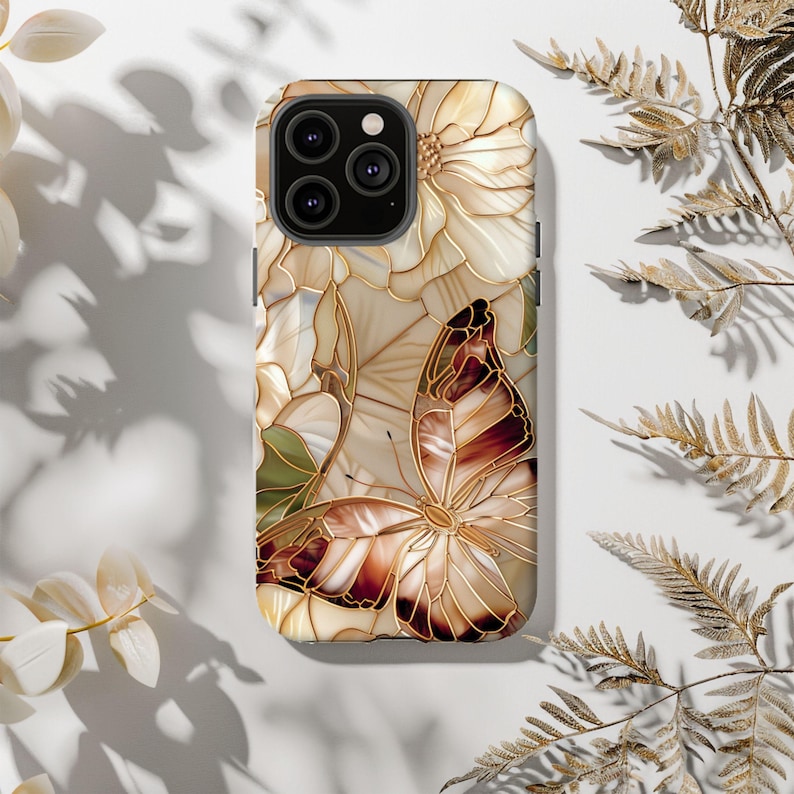 May include: A phone case featuring a floral and butterfly design. The case has a cream-colored background with a large butterfly in shades of brown and gold. Flowers and leaves are also depicted in the design, with gold outlines. The case has a glossy finish.
