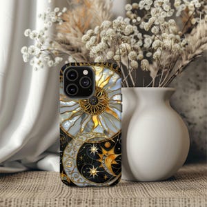 May include: A smartphone case featuring a celestial design with a sun, moon, and stars. The case has a black background with gold and white accents. The sun is gold with a white and blue center, and the moon is white with gold details.