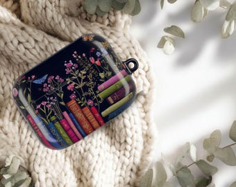 May include: A black AirPod case featuring a floral and book design. The case is adorned with colorful flowers, butterflies, and a stack of vintage books. The case has a small loop for attaching to a keychain.
