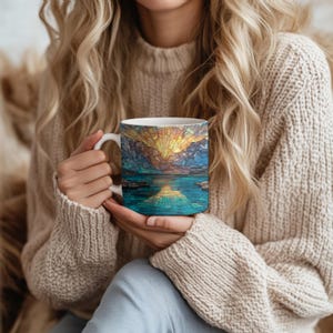 May include: White ceramic mug featuring a stained-glass design of a sunrise over a lake and mountains. The design incorporates shades of blue, yellow, and orange. The mug has a white handle and is held by a person wearing a beige sweater.