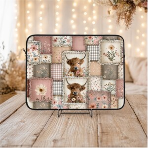 Puede incluir: Una funda para portátil con un diseño de patchwork con motivos florales y dos vacas de las Tierras Altas. La paleta de colores incluye tonos rosas, beige y marrones. La funda se muestra sobre una superficie de madera.
