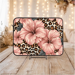 May include: A laptop sleeve featuring a floral design with large pink hibiscus flowers and gray leaves set against a leopard print background. The sleeve has a black zipper and trim, and is displayed on a wooden surface.