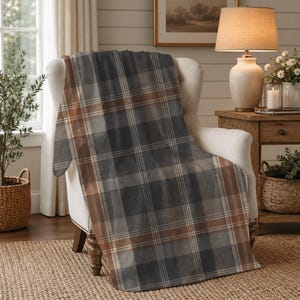 May include: A soft throw blanket featuring a classic plaid pattern in shades of charcoal gray, muted brown, and thin white intersecting lines. The fabric is draped over the back and seat of an upholstered armchair, displaying a rectangular shape.