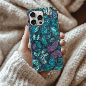 May include: A smartphone case featuring a mosaic design of butterflies in shades of teal, purple, and blue. The case has a dark blue background and a glossy finish. The case is held in a hand.