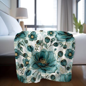 May include: A teal and gold floral throw blanket. The blanket features a repeating pattern of large teal flowers with gold outlines and accents, set against a white background. The flowers are in various stages of bloom, with detailed petals and buds.