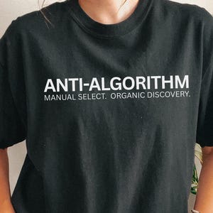 May include: Black t-shirt with the white text "ANTI-ALGORITHM" above "MANUAL SELECT. ORGANIC DISCOVERY." The shirt is size large. The person is wearing a bracelet. The background includes a macrame wall hanging and a plant.