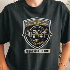 May include: A black short-sleeve t-shirt featuring a graphic shield emblem with the text FIRST RESPONDER and 911 DISPATCHER. The design includes icons of a badge, ladder, medical cross, and caduceus, with the text ANSWERING THE CALL below.