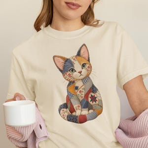 Könnte beinhalten: Ein cremefarbenes Kurzarm-T-Shirt mit einer zentralen Grafik einer sitzenden Katze, die aus verschiedenen Patchwork-Stoffmustern in Blau-, Rot-, Gelb- und Weißtönen besteht, mit sichtbaren Nahtdetails am gesamten Körper der Katze.
