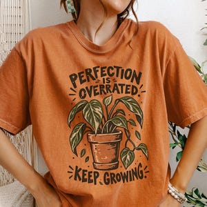 May include: A burnt orange short-sleeve t-shirt featuring a graphic of a potted plant with green leaves. The text PERFECTION IS OVERRATED and KEEP GROWING is printed in black above and below the plant illustration. The shirt is labeled YAM Size Large.