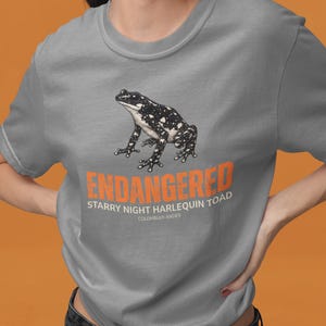 May include: A grey crew neck t-shirt featuring a graphic illustration of a black and white spotted toad. Below the toad, the text reads ENDANGERED in orange, followed by STARRY NIGHT HARLEQUIN TOAD and COLOMBIAN ANDES in white lettering.