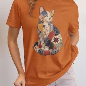 Könnte beinhalten: Ein rostorangefarbenes Kurzarm-T-Shirt mit einer zentralen Grafik einer sitzenden Katze, die aus verschiedenen Patchwork-Stoffmustern zusammengesetzt ist, darunter Blumen-, Vichy- und Sternmotive in Blau-, Rot-, Creme- und Brauntönen.