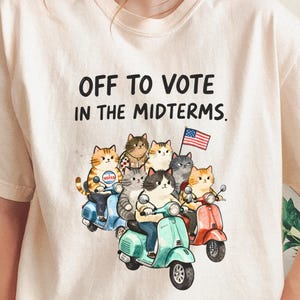 May include: An ivory t-shirt featuring the black text OFF TO VOTE IN THE MIDTERMS. Below the text is a colorful illustration of seven cats riding three scooters, one of which is blue, one teal, and one red, with a small American flag attached.