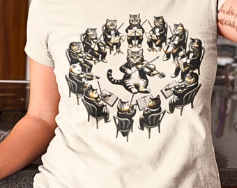 May include: Cream-colored t-shirt featuring a whimsical illustration of a cat orchestra. The cats, dressed in formal attire, are arranged in a circle, playing various instruments. A unique and artistic design for cat lovers.