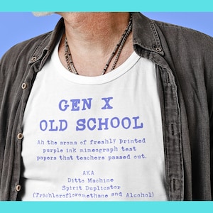 May include: White t-shirt with the text "GEN X OLD SCHOOL" in blue. The shirt also includes additional text about mimeograph test papers and the term "Ditto Machine". The shirt is worn under a gray button-up shirt.