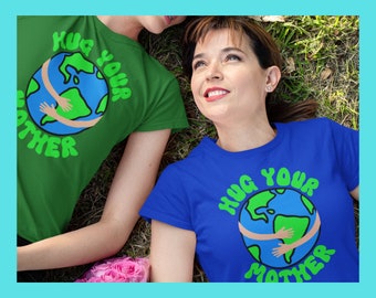 May include: Two people wearing green and blue t-shirts with the text "Hug Your Mother" and a graphic of a globe with hands around it. The globe is green and blue with brown continents. The people are lying on green grass with pink flowers.