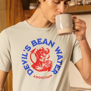May include: A light gray t-shirt with a blue and red graphic of a devil holding a cup of coffee. The graphic is in a circle with the text "Devil's Bean Water Addiction".