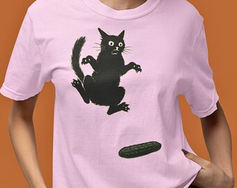 Può includere: T-shirt rosa chiaro con una silhouette nera di un gatto spaventato che salta via da un cetriolo verde. Il gatto ha grandi occhi e zampe tese. La t-shirt è uno stile girocollo.