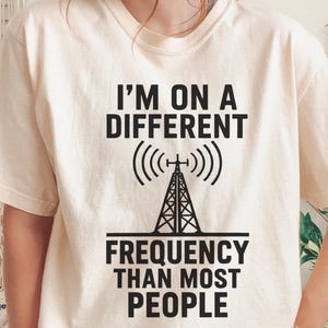 Peut inclure: T-shirt de couleur ivoire avec l'inscription noire "I'M ON A DIFFERENT FREQUENCY THAN MOST PEOPLE". Le motif comprend une antenne radio avec des ondes. Le t-shirt est de taille large.