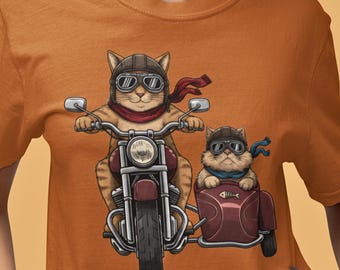 Puede incluir: Camiseta naranja con una ilustración de dibujos animados de dos gatos en una motocicleta. Un gato conduce la moto, el otro está en un sidecar. Ambos gatos llevan cascos y bufandas.