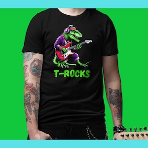 Puede incluir: Una camiseta negra con un dinosaurio verde que lleva auriculares y toca la guitarra. El dinosaurio lleva una chaqueta morada y un pañuelo morado en la cabeza. El texto "T-ROCKS" está impreso en verde debajo del dinosaurio.