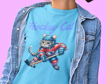 Op de afbeelding: Lichtblauw T-shirt met een cartoonkat die hockey speelt. De kat draagt een helm en een rood-blauw shirt en houdt een hockeystick vast. De tekst "Hockey Cat" staat in lichtpaarse kleur boven de kat.