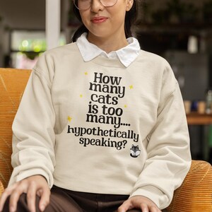 Puede incluir: Sudadera color crema con el texto negro "How many cats is too many... hypothetically speaking?". Se usa con una camisa blanca con cuello. La persona está sentada en una silla naranja.