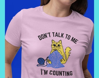May include: Pink t-shirt with a yellow cat knitting a blue scarf and the text "Don't talk to me I'm counting".