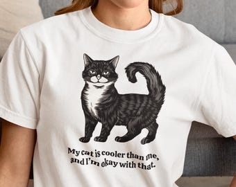 May include: White t-shirt with a black and white illustration of a cat. The text "My cat is cooler than me, and I'm okay with that." is printed below the cat.