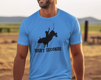 May include: A man wearing a light blue t-shirt with a black silhouette of a cowboy riding a bull and the text "Eight Seconds". He is also wearing a brown cowboy hat and blue jeans.