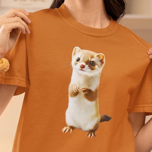 May include: An orange t-shirt with a cartoon ferret design. The ferret is shown standing upright with its paws clasped. It has a white body, brown patches, and a black tail. The t-shirt is a plain orange colour.