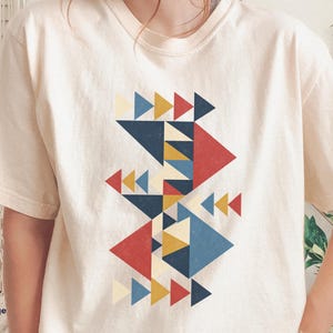Puede incluir: Una camiseta de manga corta color marfil con un diseño gráfico central compuesto por varios triángulos geométricos en tonos azul marino, amarillo mostaza, rojo y crema. El texto "IVORY" y "Size Large" aparece en la esquina inferior izquierda.