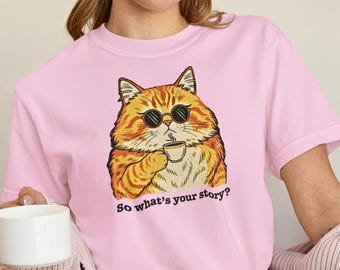 May include: Pink t-shirt featuring a cartoon cat wearing sunglasses and holding a coffee cup. The cat is orange and white with black sunglasses. The text "So what's your story?" is printed below the cat.