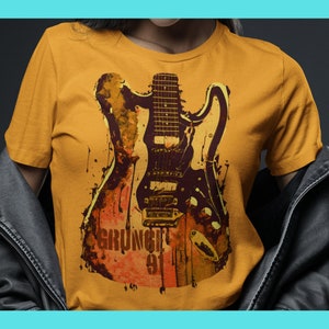 Puede incluir: Una camiseta amarilla con un estampado gráfico de una guitarra eléctrica negra con un diseño de estilo grunge. El texto "GRUNGE 91" está impreso en la camiseta.