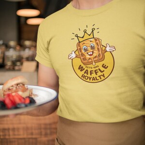 May include: Yellow t-shirt featuring a cartoon waffle wearing a crown, with the text "WAFFLE ROYALTY ESTD 1890" in a circular design. The waffle has a smiling face and syrup dripping down. The shirt is worn by a person.