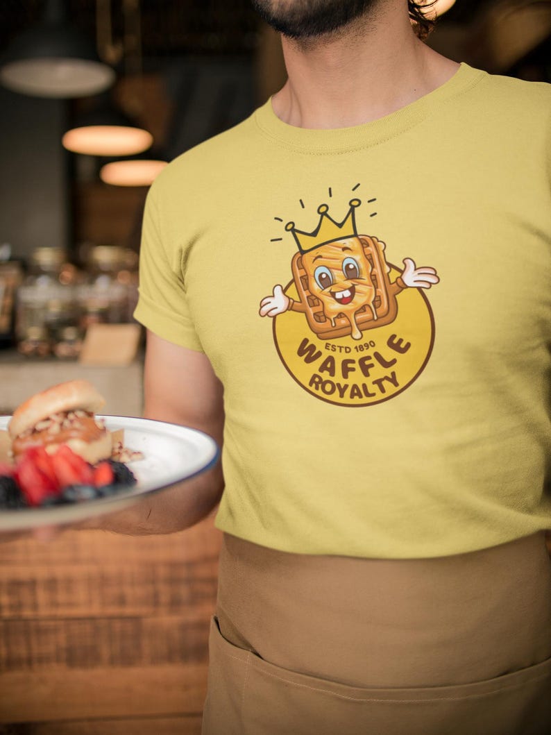 May include: Yellow t-shirt featuring a cartoon waffle wearing a crown, with the text "WAFFLE ROYALTY ESTD 1890" in a circular design. The waffle has a smiling face and syrup dripping down. The shirt is worn by a person.