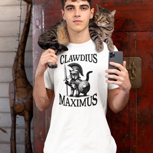 Könnte beinhalten: Weißes T-Shirt mit einer Schwarzweißillustration einer Katze, die als römischer Soldat verkleidet ist. Der Text "CLAUDIUS MAXIMUS" ist auf das Shirt gedruckt.
