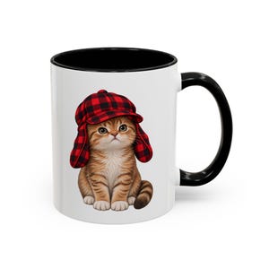 May include: A white ceramic mug with a black interior and handle, featuring a graphic of a ginger tabby kitten wearing a red and black buffalo plaid trapper hat. The kitten is sitting upright, looking forward with large eyes and white paws.